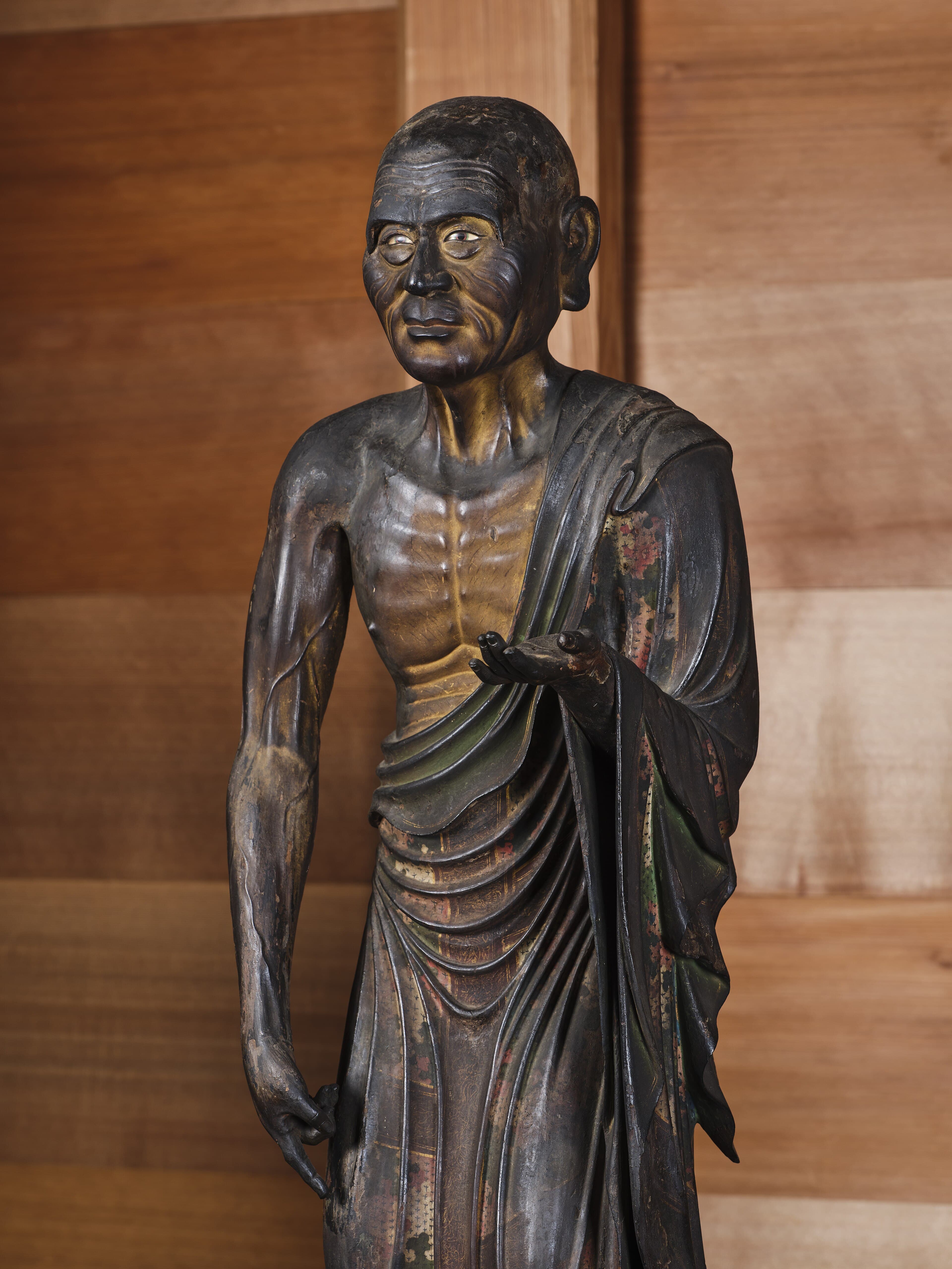 Weathered wooden statue of the elder disciple Mahamoggallana, gaunt and lined, standing in monastic robes with one hand extended