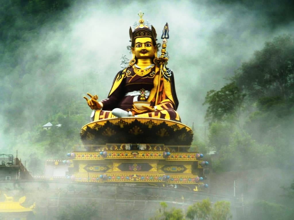 Large golden statue of Padmasambhava (Guru Rinpoche) enthroned on a lotus, crowned and holding a vajra and skull cup, rising above mist-covered hills
