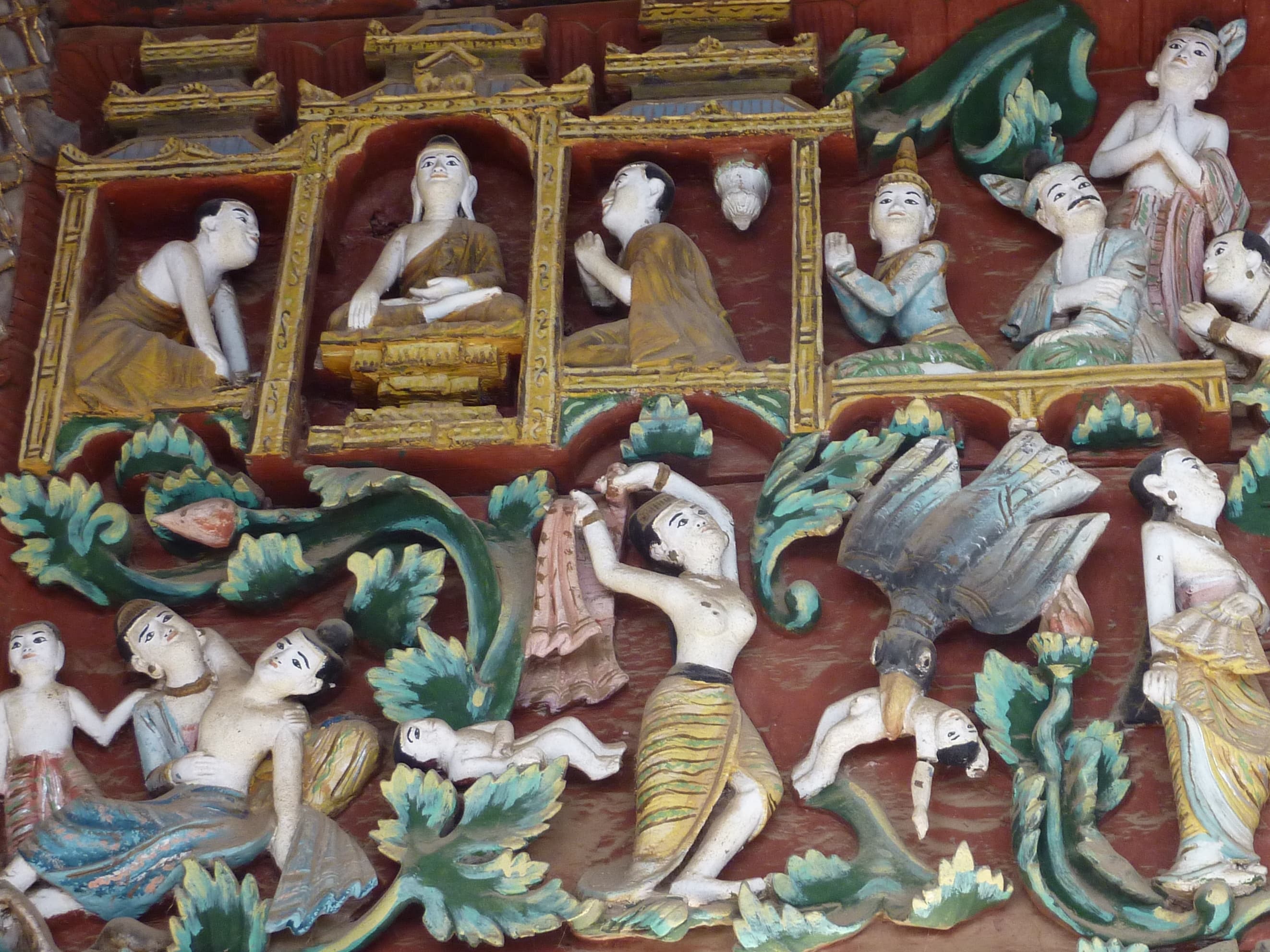 Carved and painted temple relief depicting the tragic narrative of Patacara — figures with infants amid floodwaters, a bird carrying a child, beneath a shrine of seated Buddha images